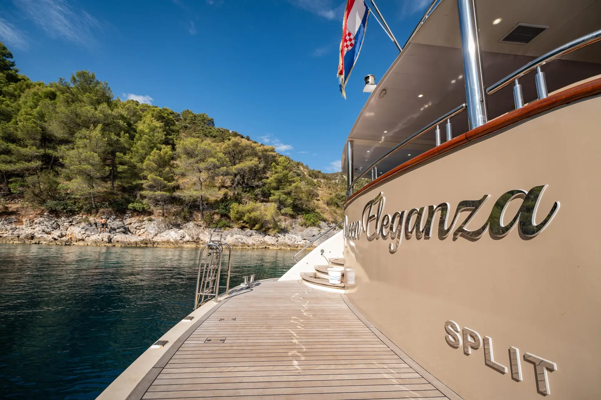 A yacht named Eleganza docked near a rocky, tree-covered shoreline, with a clear blue sky and calm water. The name Eleganza and Split are visible on the yachts side.