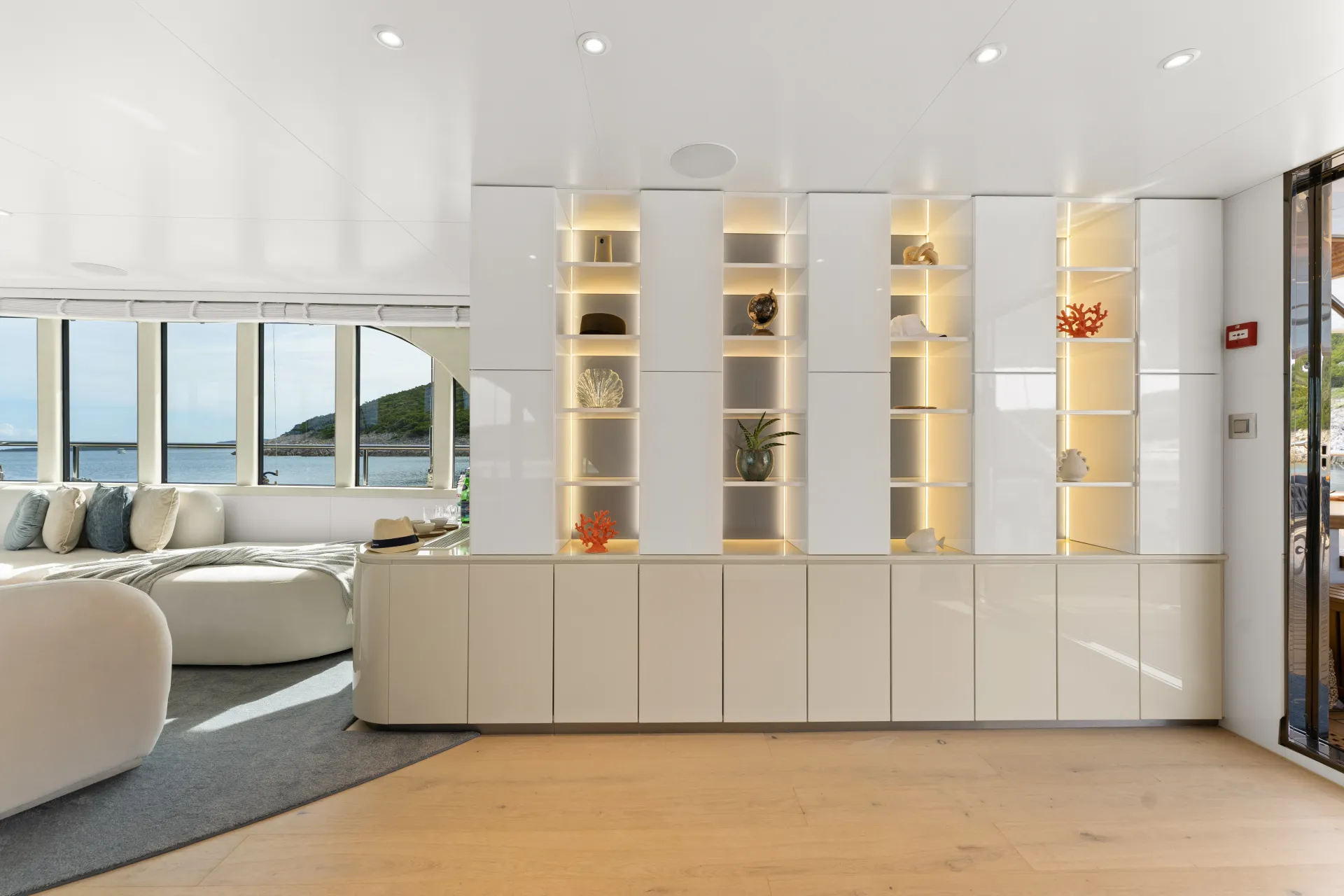 MAXITA Modern, bright living space with built-in white shelves displaying decorative items and soft lighting. Large windows offer a scenic view of the ocean and greenery outside. Contemporary white sofas sit on a gray rug.