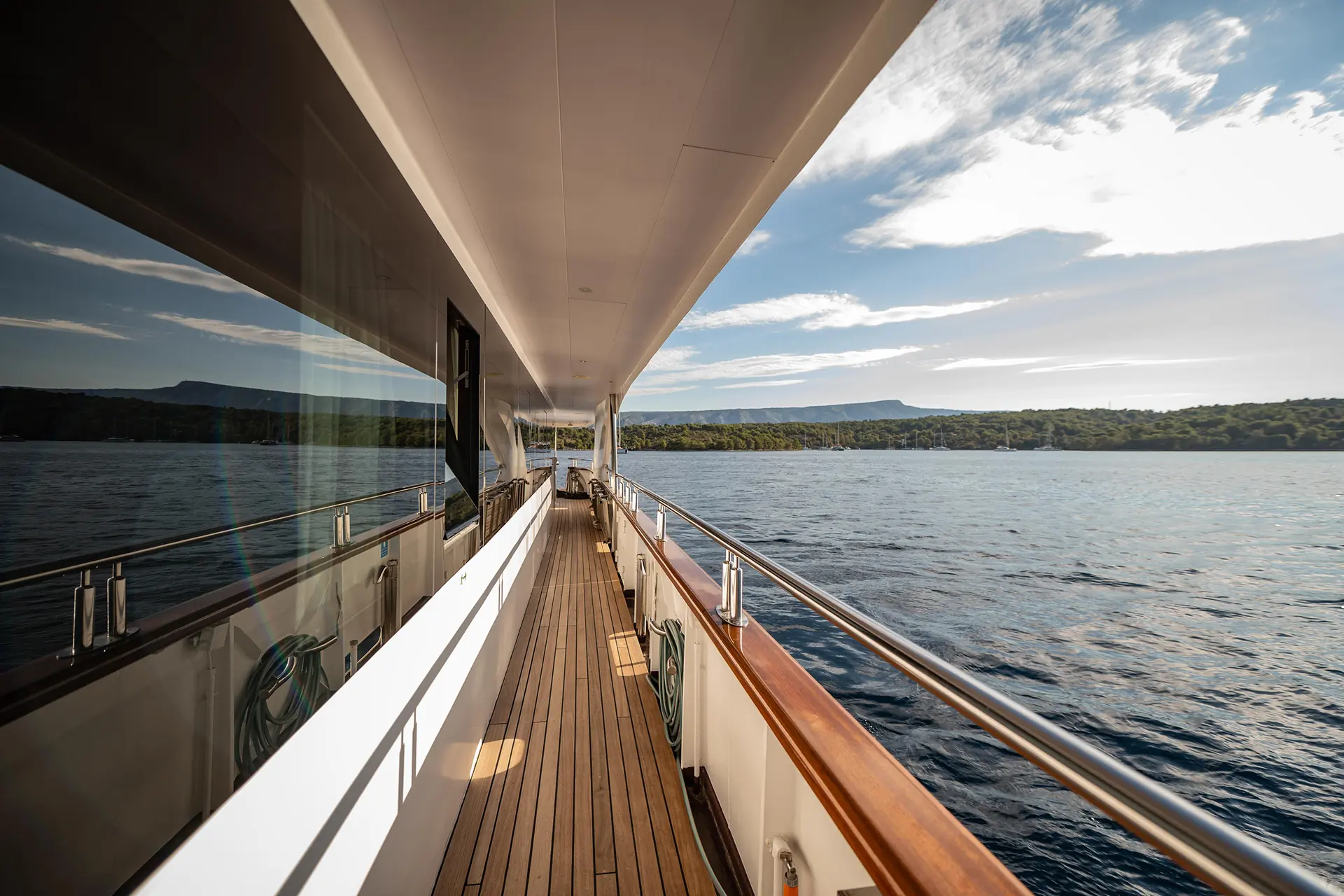 A wooden deck walkway runs along the side of a yacht, overlooking calm blue water and a green shoreline under a partly cloudy sky.