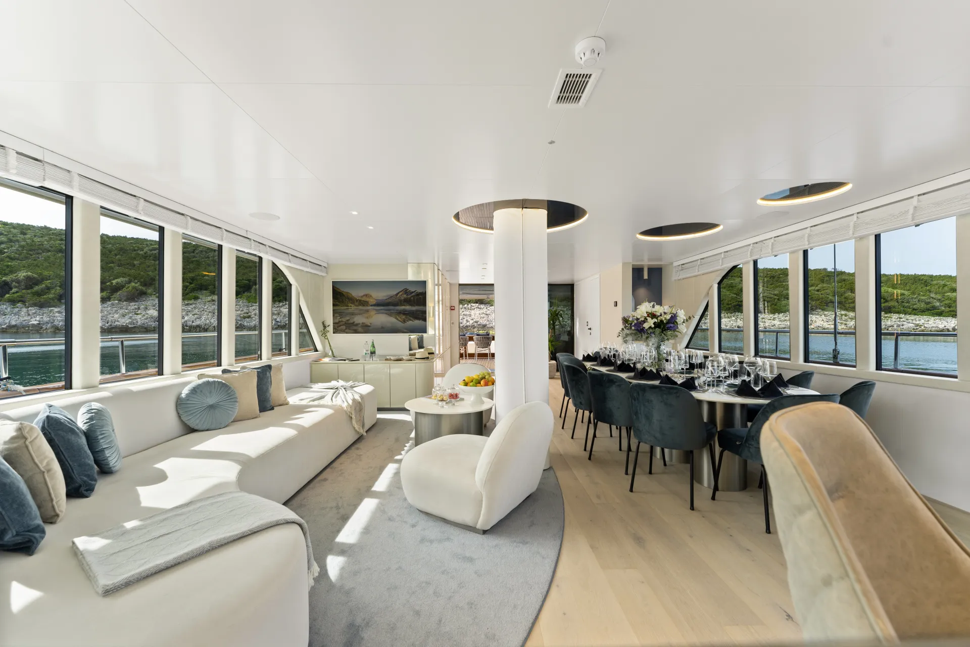 MAXITA Modern yacht interior with large windows, white curved sofa, round chairs, dining table set for eight, and scenic views of water and greenery outside. Natural light fills the elegant, open living and dining area.