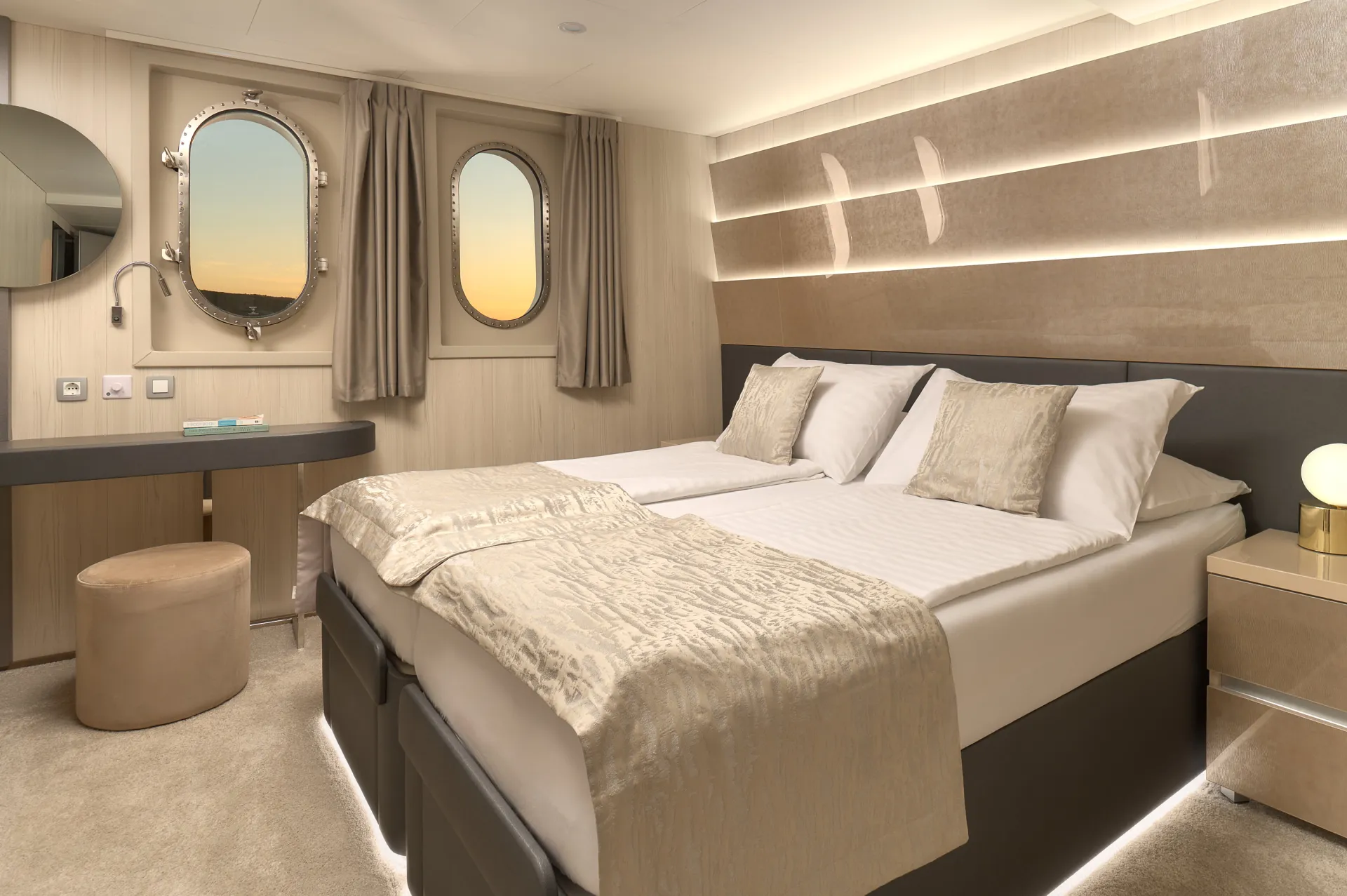 CRISTAL Modern cruise ship cabin with two beds pushed together, neutral-toned bedding, two round windows with beige curtains, a vanity desk with a stool, and soft ambient lighting.