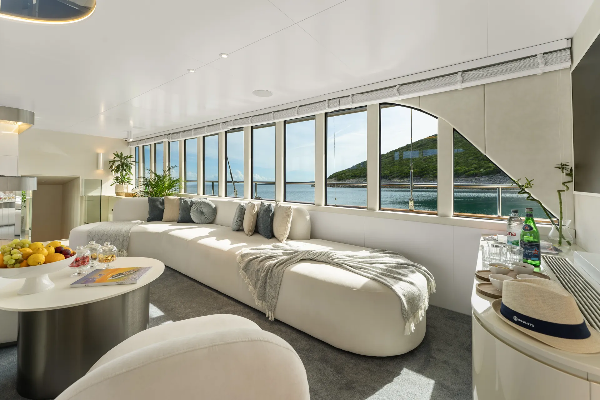MAXITA Modern yacht interior with large windows, a long curved beige sofa with cushions and a throw, a round white table with fruit, and a view of the sea and green hills outside. Light and airy, with natural decor.