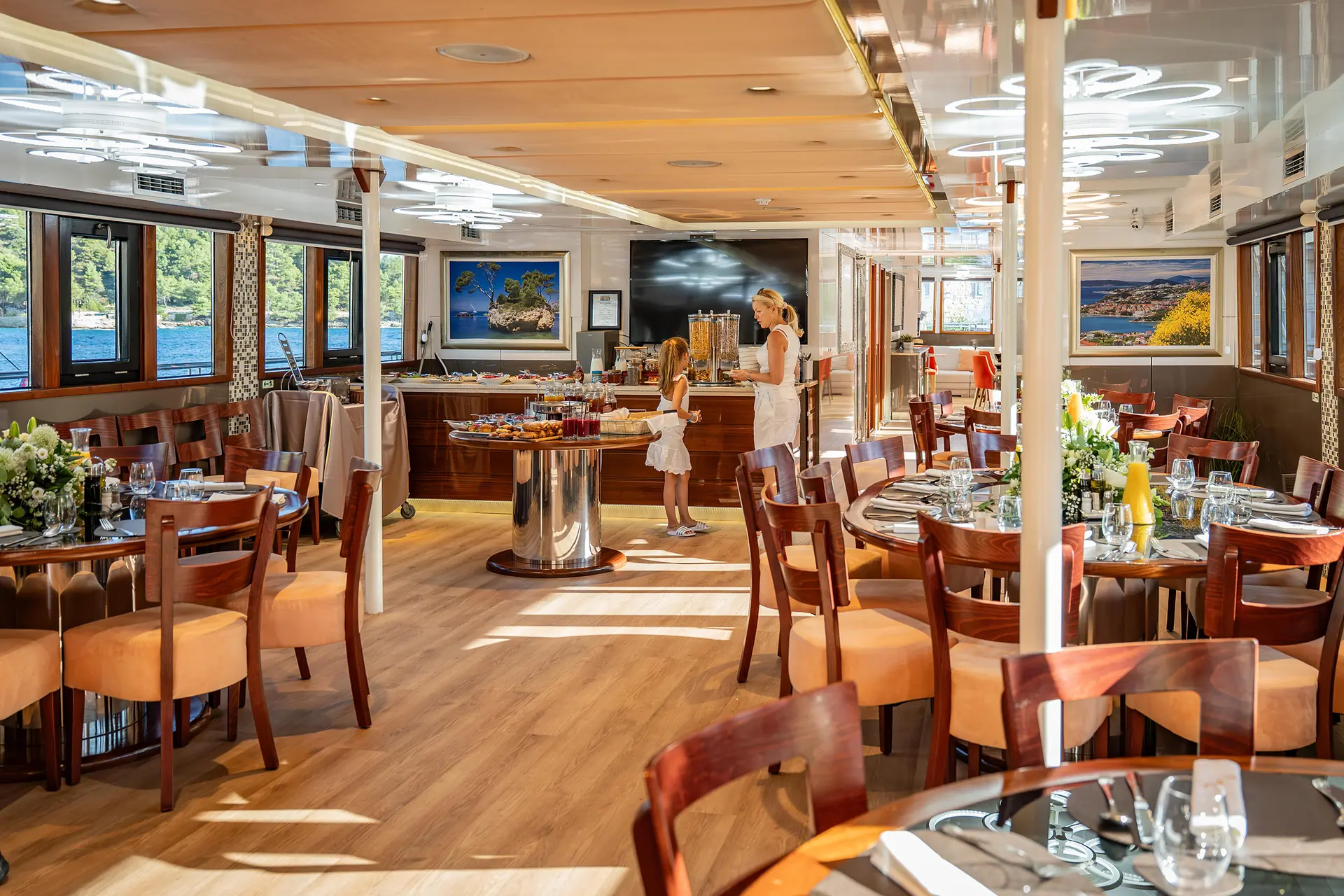 Spacious yacht dining room with wooden floors, elegant tables set for meals, buffet area, and large windows. Two people are near the buffet; sunlight fills the space.