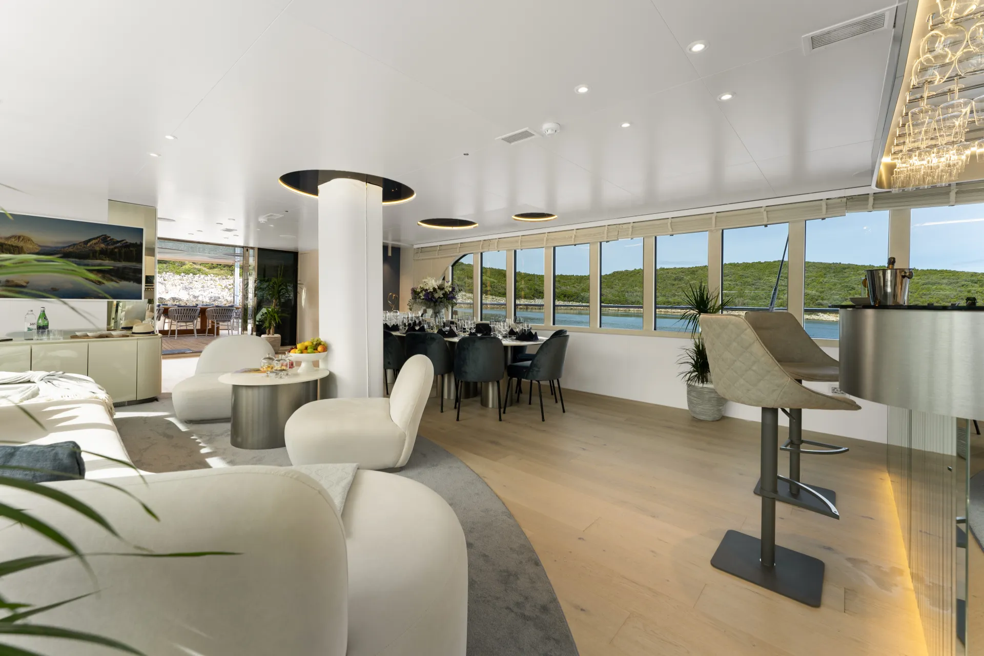 MAXITA Luxurious yacht interior with white sofas, a dining table set for a meal, modern decor, large windows offering scenic water and green hillside views, and a bar area with stools. Natural light fills the space.