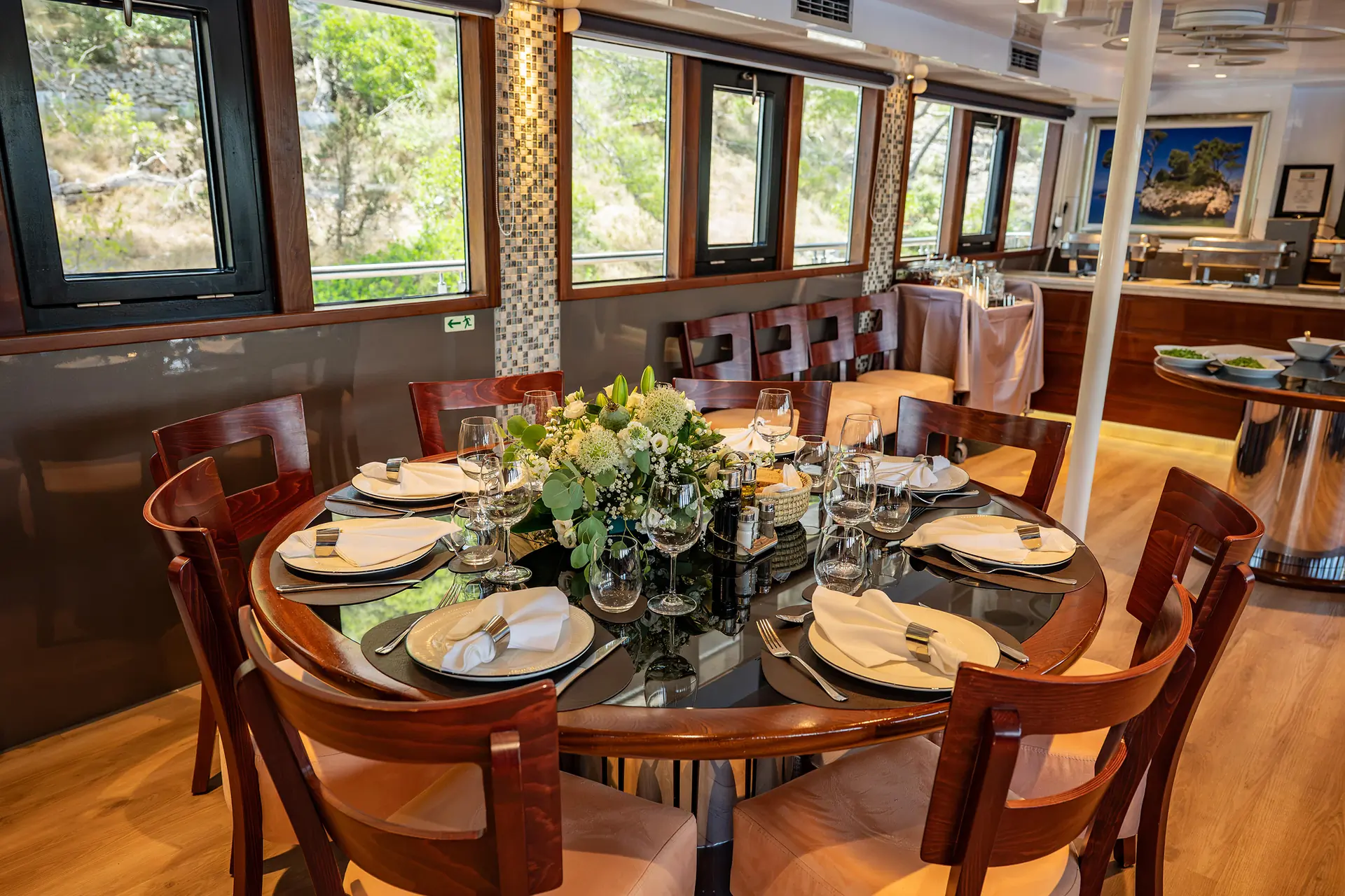 A round dining table with eight wooden chairs is set with white plates, napkins, glassware, and a floral centerpiece in a bright, modern room with large windows and wooden floors.