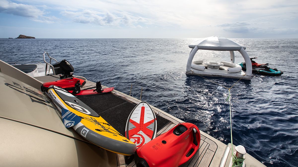 YCM 120 A yacht deck with paddleboards, a jet ski, and water gear overlooks calm blue sea. Nearby, a white inflatable floating lounge sits on the water under a cloudy sky.