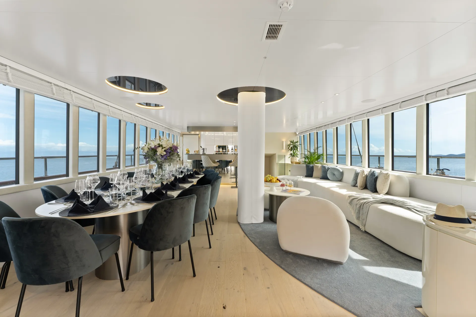 MAXITA A modern yacht interior with large windows, elegant dining tables set with glassware and black napkins, curved white sofas, decorative plants, and a view of the ocean outside. Natural light fills the spacious room.