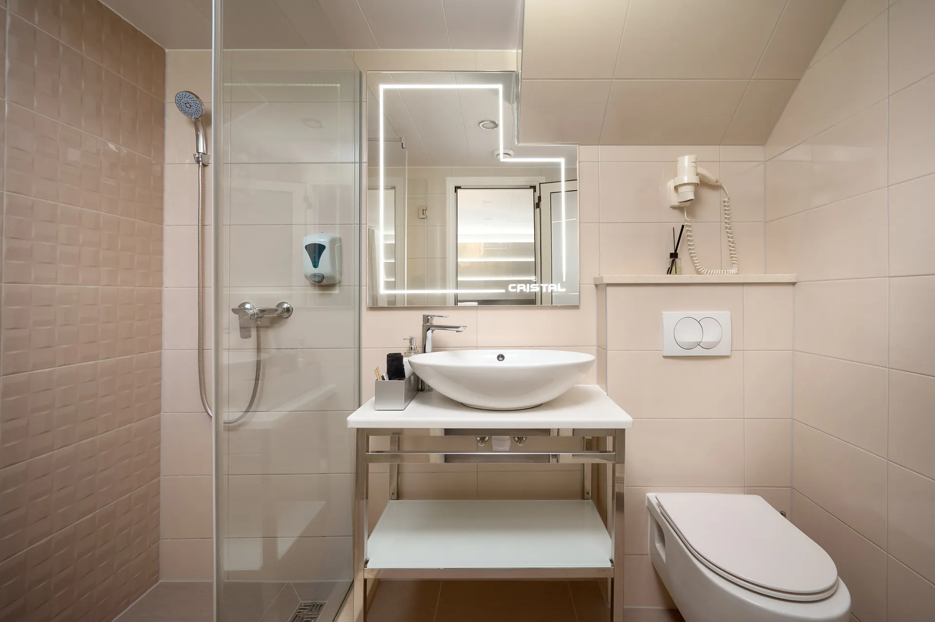 CRISTAL Modern bathroom with glass shower, wall-mounted showerhead, floating sink with large mirror, hairdryer, soap dispenser, and wall-mounted toilet. Neutral beige tiles and clean, minimalist design.