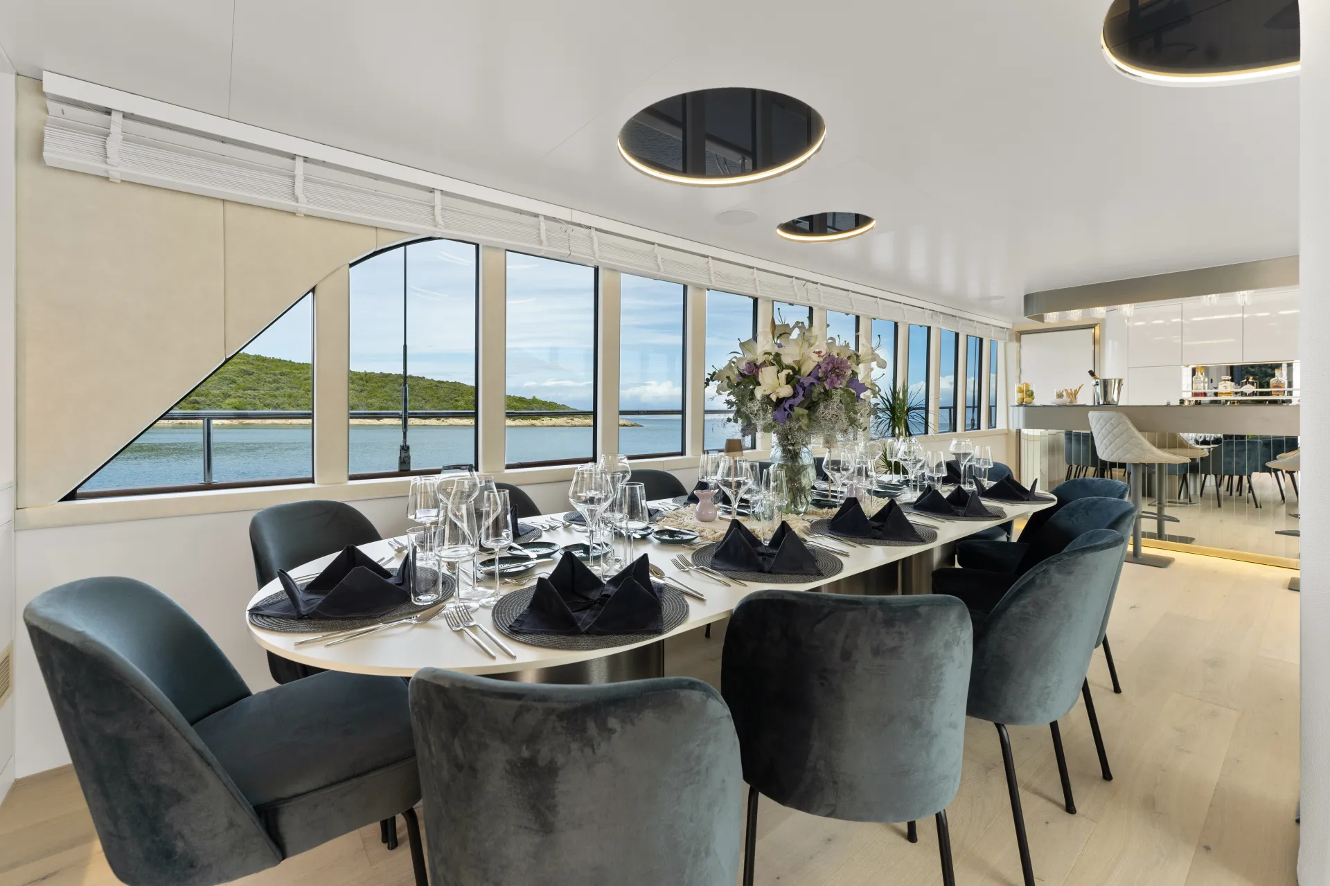 MAXITA A modern dining area with a large oval table set for eight, featuring black napkins and glassware. Plush blue chairs surround the table. Large windows offer a scenic view of water and greenery outside.