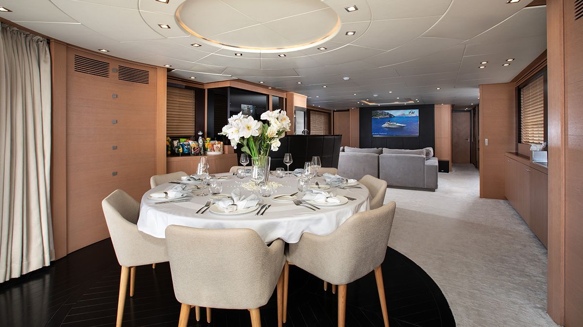 YCM 120 A modern dining area on a yacht with a round table set for six, beige chairs, a flower centerpiece, and a view of a cozy lounge with a large TV displaying a yacht on the water.