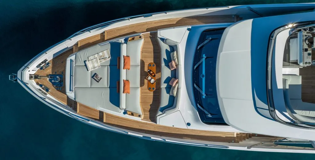 VIVA II Aerial view of a luxurious yachts bow deck with cushioned seating, a table set with drinks, open magazines, and decorative pillows, floating on calm, clear blue water.