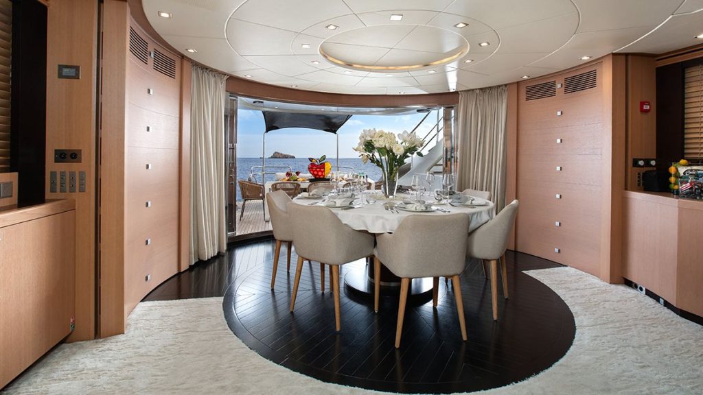 YCM 120 Elegant yacht dining area with a round table set for six, surrounded by light wood walls, large windows, and doors opening to a deck with a sea view. A bouquet of white flowers is at the center of the table.
