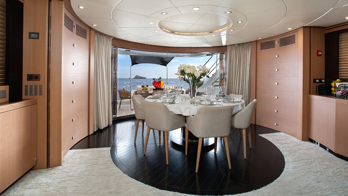 YCM 120 Elegant yacht dining area with a round table set for six, surrounded by light wood walls, large windows, and doors opening to a deck with a sea view. A bouquet of white flowers is at the center of the table.