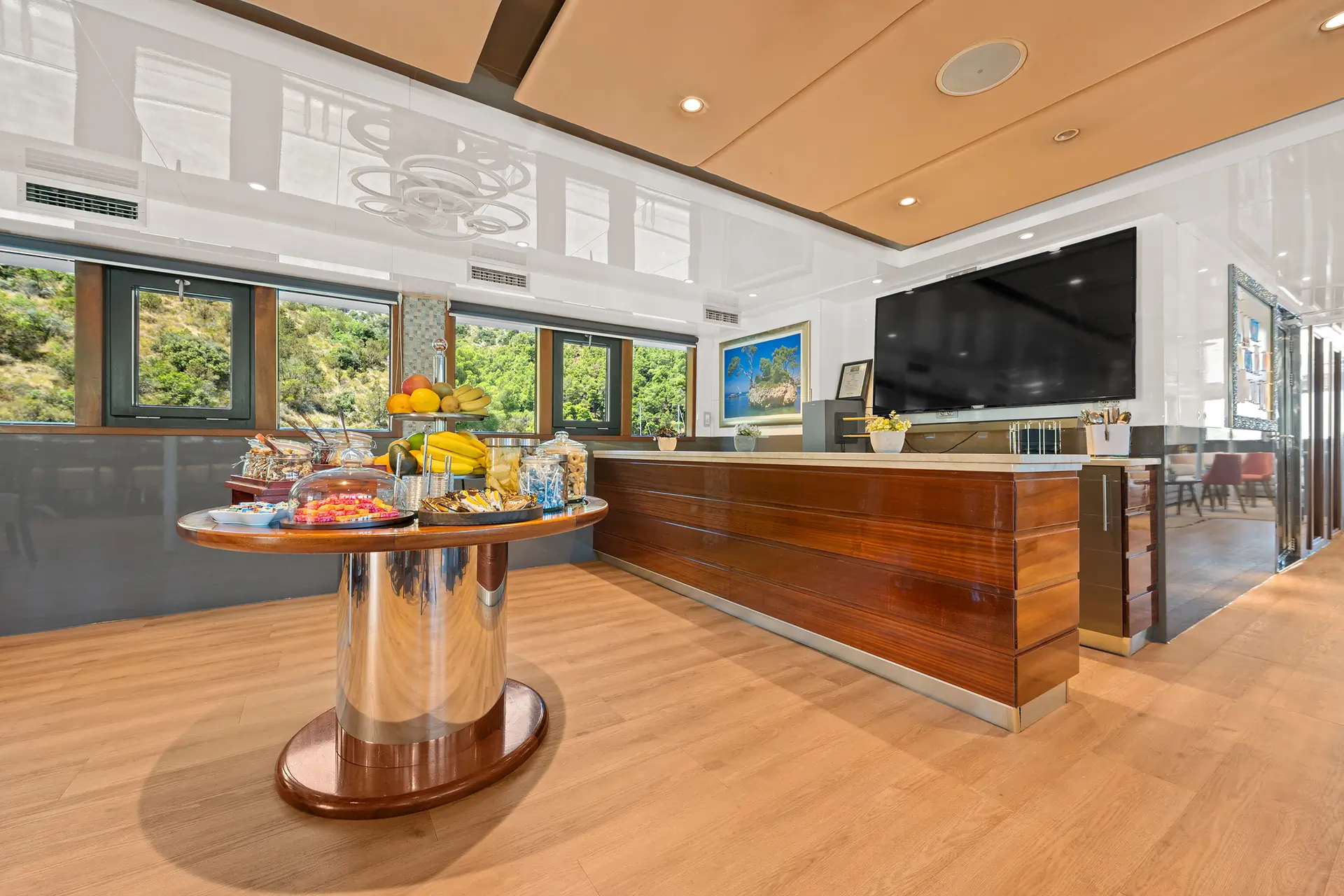 A modern, bright room with wooden floors, large windows, a wooden reception desk, a wall-mounted TV, and a round table with snacks, fruit, and drinks. Lush greenery is visible outside.