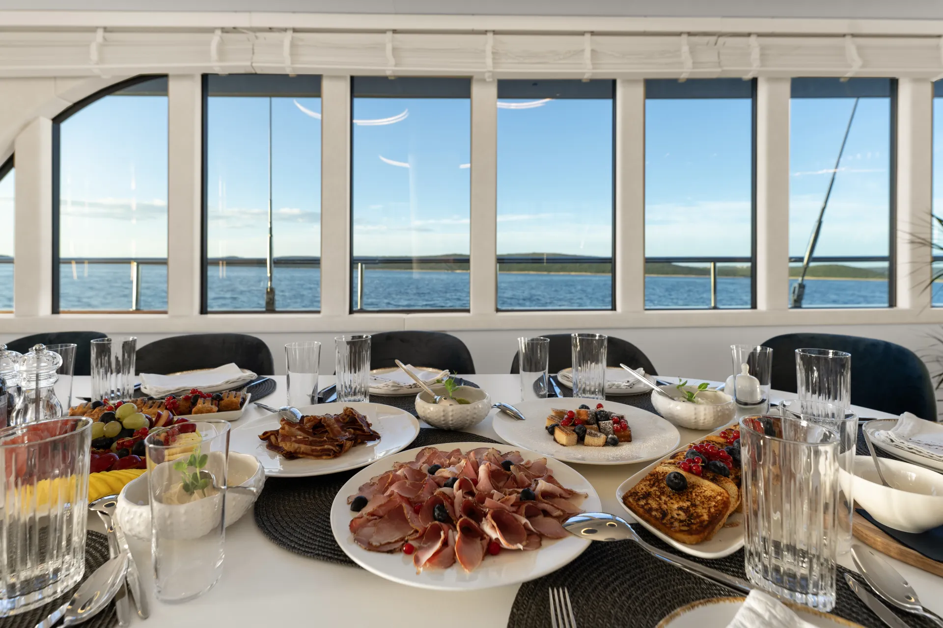 MAXITA A dining table set with plates of assorted meats, fruit, pastries, and glasses, positioned next to wide windows offering a scenic view of the sea and distant hills under a clear sky.