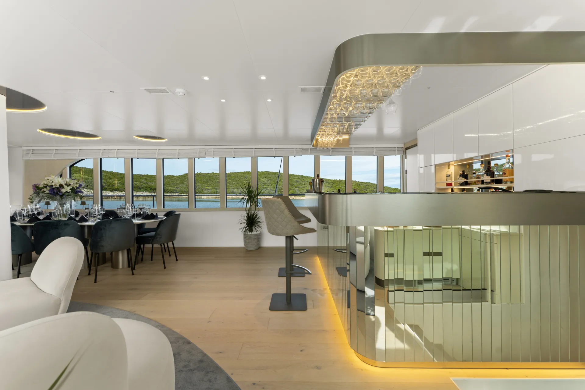 MAXITA Modern yacht interior featuring a stylish bar with glassware, stools, a dining area set for guests, large windows with scenic water and greenery views, and elegant contemporary decor.