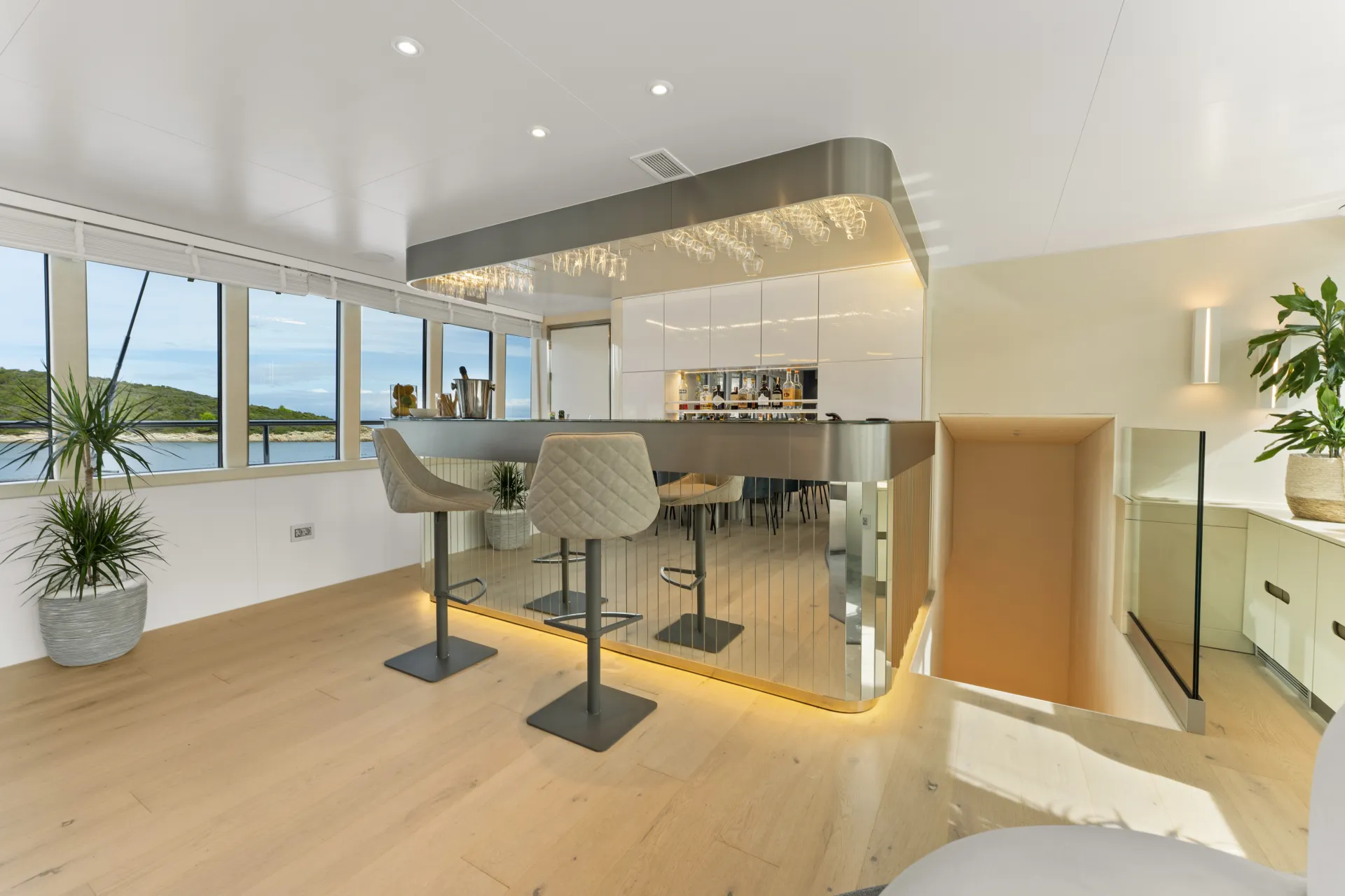 MAXITA A modern, stylish bar area with two quilted bar stools, mirrored bar front, and overhead glass rack. Large windows offer scenic water views, and potted plants add greenery to the light wood and white interior.