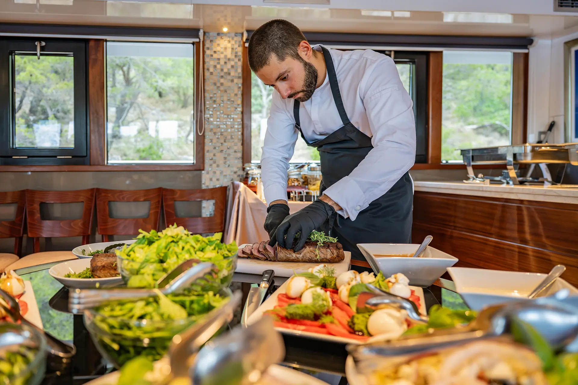A chef in a white shirt and black apron slices meat in a restaurant kitchen, surrounded by plates of fresh vegetables and salads on a counter.