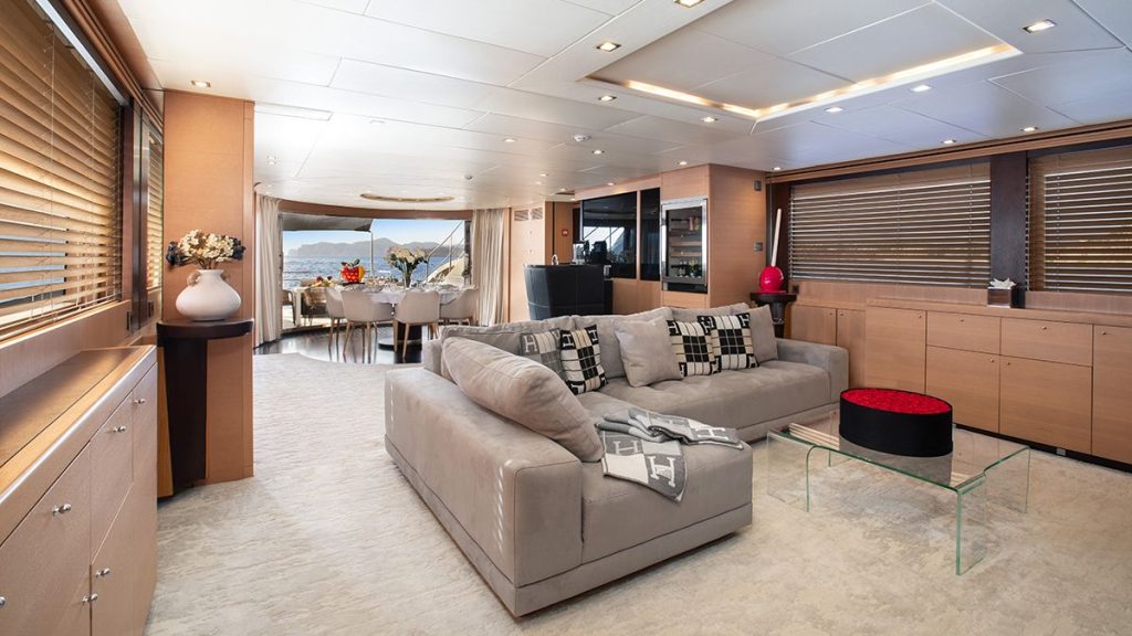 YCM 120 Luxurious yacht interior with a spacious living area featuring a large gray sectional sofa, modern decor, wood paneling, and large windows. Dining area and view of the ocean are visible in the background.