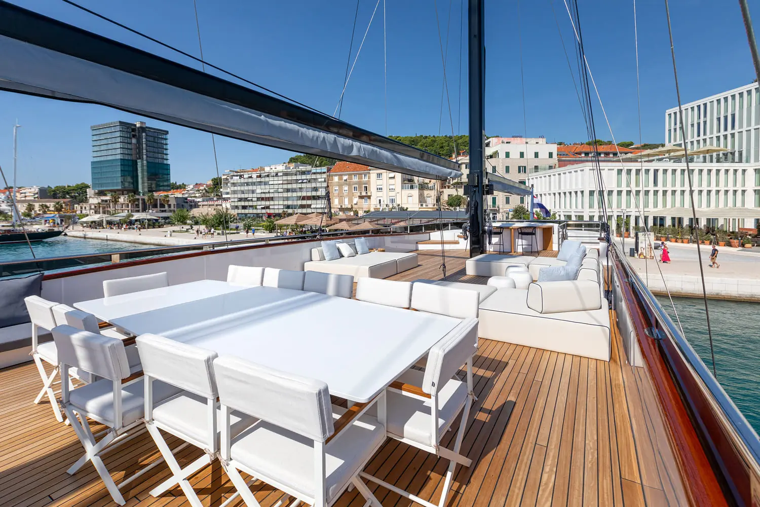 A luxury yacht deck with white dining tables, chairs, and cushioned lounge seating. Modern buildings and a marina are visible in the background under a clear blue sky.