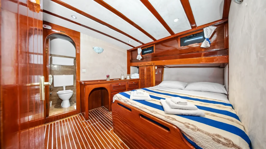 ELEGANZA A cozy boat cabin with a double bed, blue and white striped bedding, folded towels, wooden furnishings, and an en-suite bathroom visible through an open door. The cabin features ceiling beams and small windows.