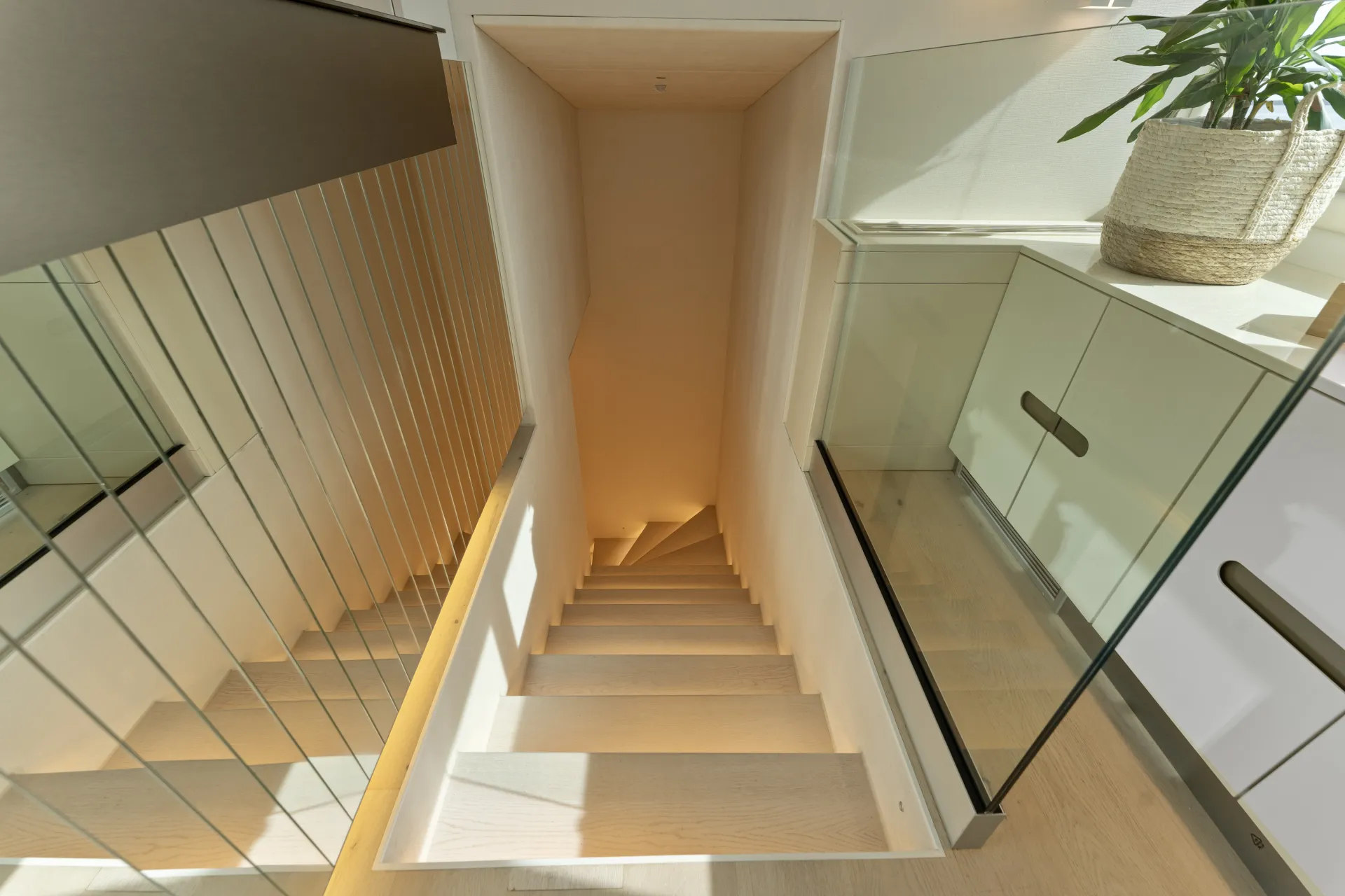MAXITA A narrow, steep wooden staircase leads downward between mirrored and glass walls in a bright, modern interior with a potted plant on a cabinet nearby.