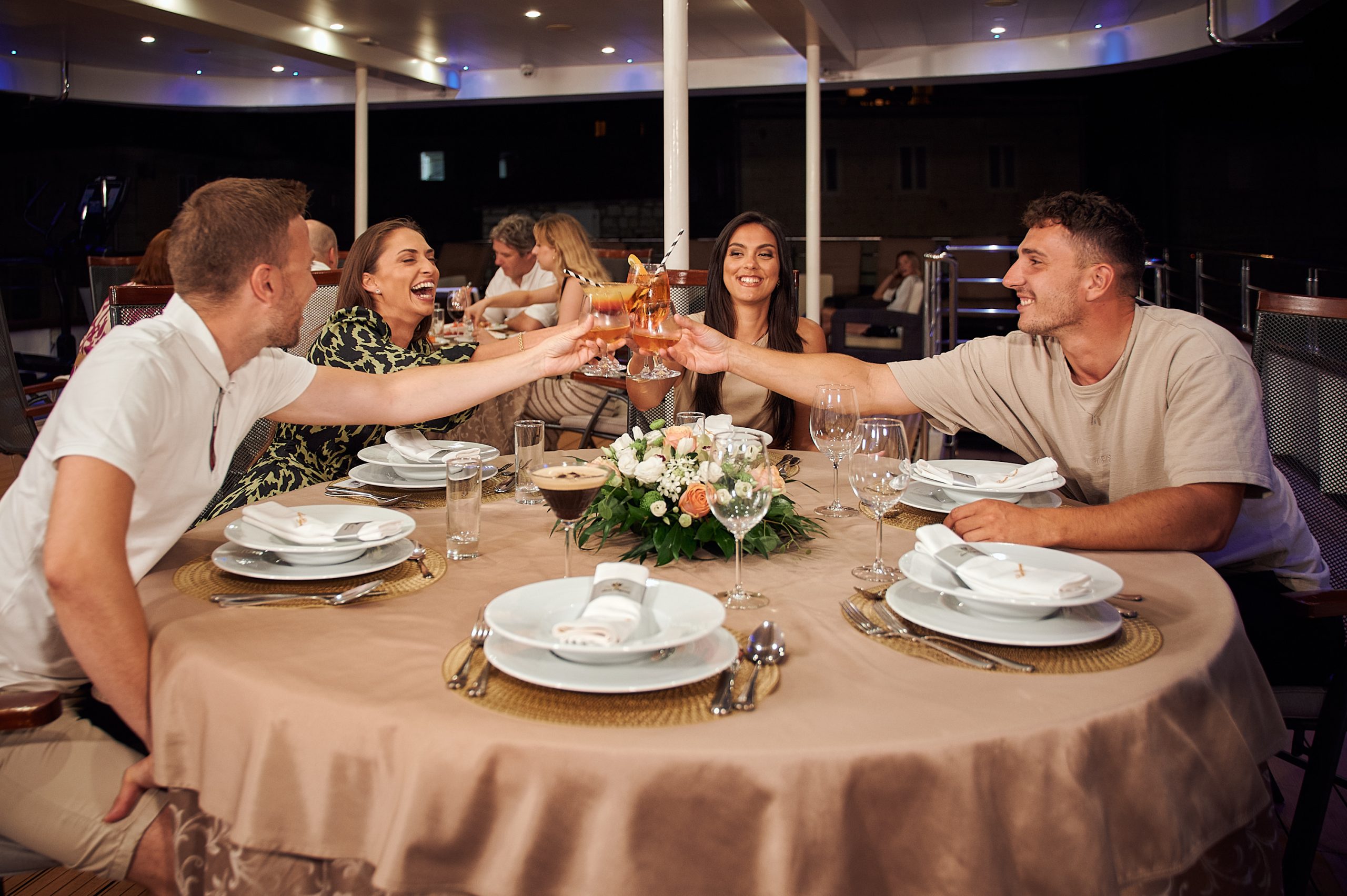 Four people sit around a round table set for dinner on a boat, smiling and raising glasses in a toast. The table is decorated with a floral centerpiece and elegant dinnerware. It is nighttime.