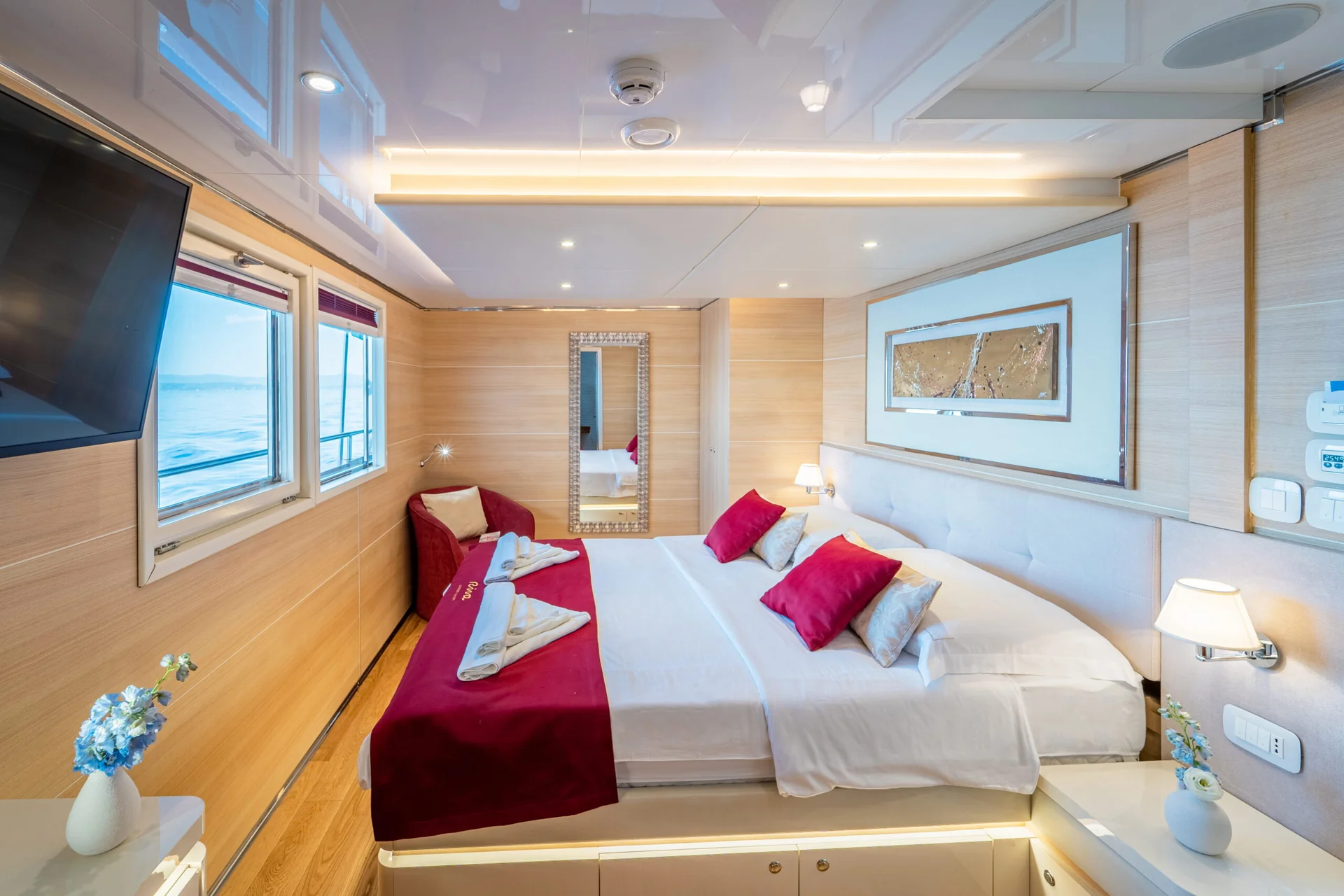 A modern yacht cabin featuring a double bed with a red runner, white linens, red throw pillows, a small armchair, a mirror, and views of the sea through the windows.