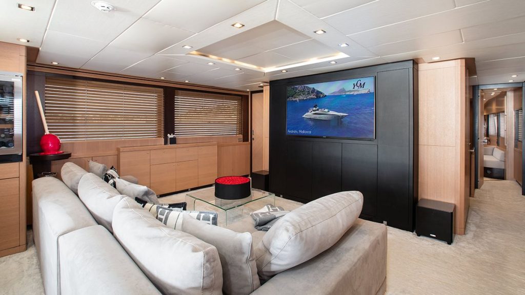 YCM 120 Modern yacht interior with a spacious lounge area, light-colored sectional sofa, decorative pillows, round ottoman, wooden paneling, large TV displaying a boat scene, and windows with horizontal blinds.