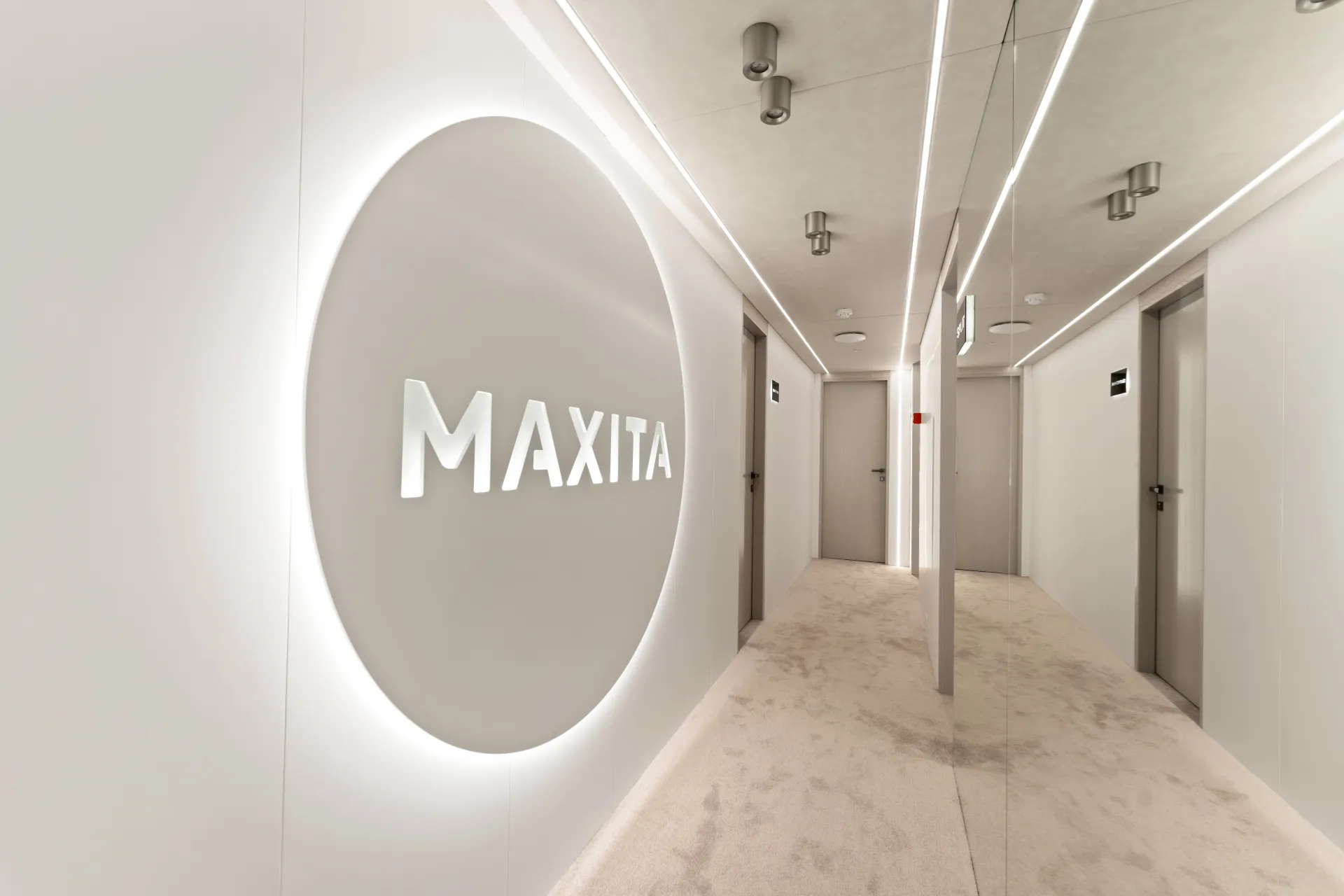 MAXITA A modern, minimalist hallway with white walls, ceiling, and carpet. The word MAXITA is illuminated on a circular wall feature. Several doors and mirrored panels line the corridor.