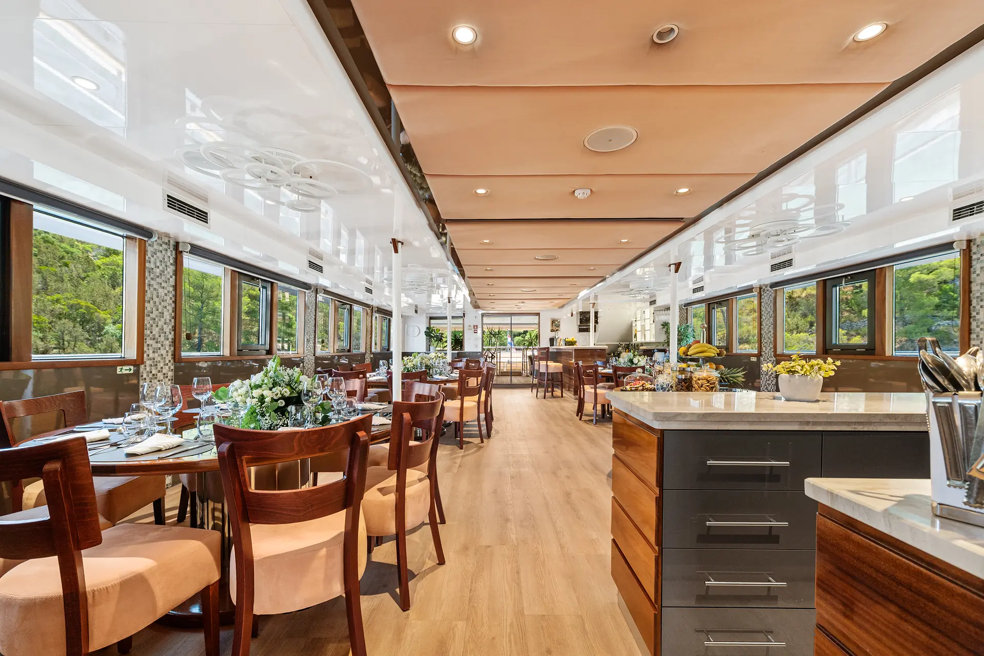 A spacious, elegant dining area with wooden tables, chairs, floral centerpieces, and large windows, offering views of greenery outside. The space is bright with natural light and modern decor.