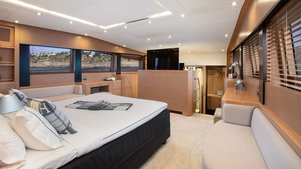YCM 120 Modern yacht bedroom with a large bed, light wood paneling, a small sofa, built-in desk, and windows with wooden blinds. The room features elegant decor and soft, ambient lighting.