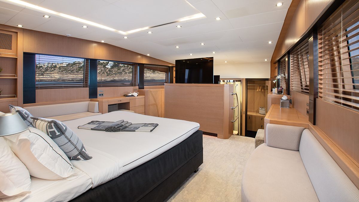 YCM 120 Modern yacht bedroom with a large bed, light wood paneling, a small sofa, built-in desk, and windows with wooden blinds. The room features elegant decor and soft, ambient lighting.