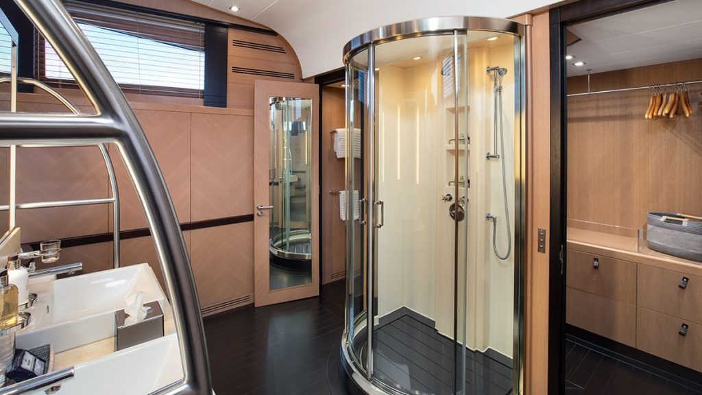 YCM 120 Modern bathroom with wood paneling, a round glass-enclosed shower, double sinks, a wall mirror, towel racks, and an open closet with hangers and a basket. Large windows provide natural light.