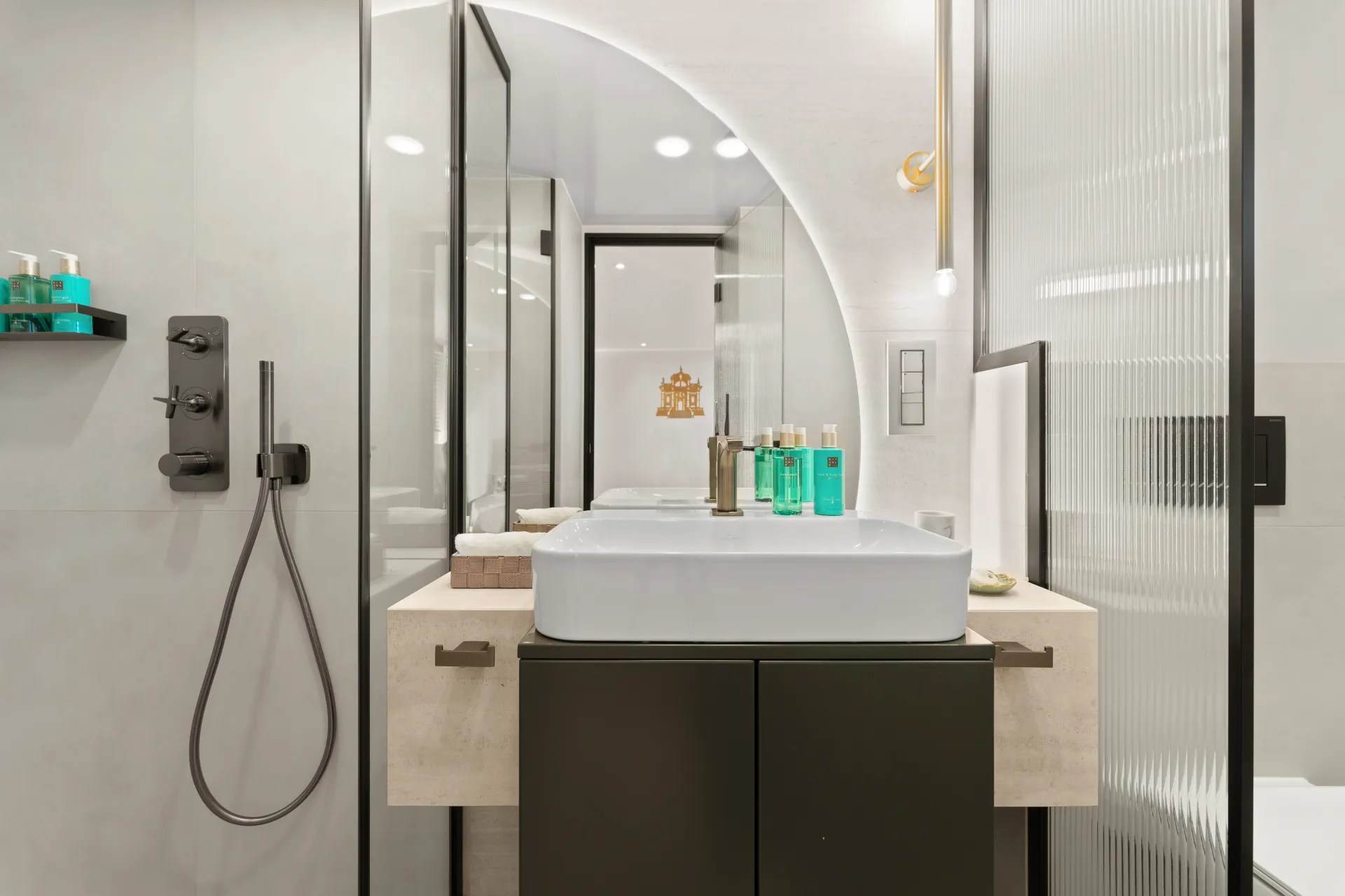 MAXITA Modern bathroom with a square white sink, black cabinet, wall-mounted shower, toiletries on the counter, mirrors, and bright lighting. Neutral tones and glass partitions create a clean, contemporary look.