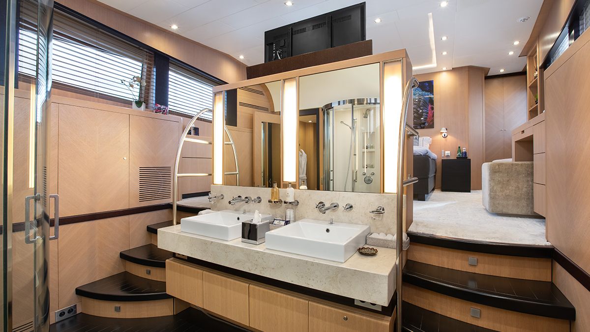 YCM 120 A modern, luxurious bathroom features dual square sinks with a large mirror, wooden accents, a glass shower, and steps leading to a spacious bedroom area with a bed and soft lighting.