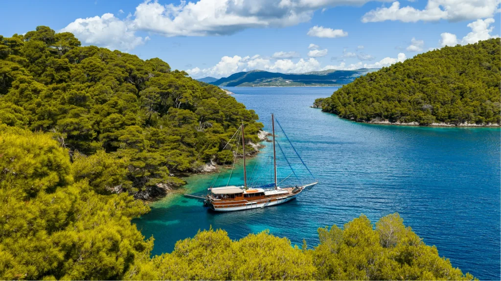 ELEGANZA A sailboat is anchored in clear blue water between two lush, green, tree-covered hills under a partly cloudy sky. The landscape in the background shows distant mountains and more greenery.