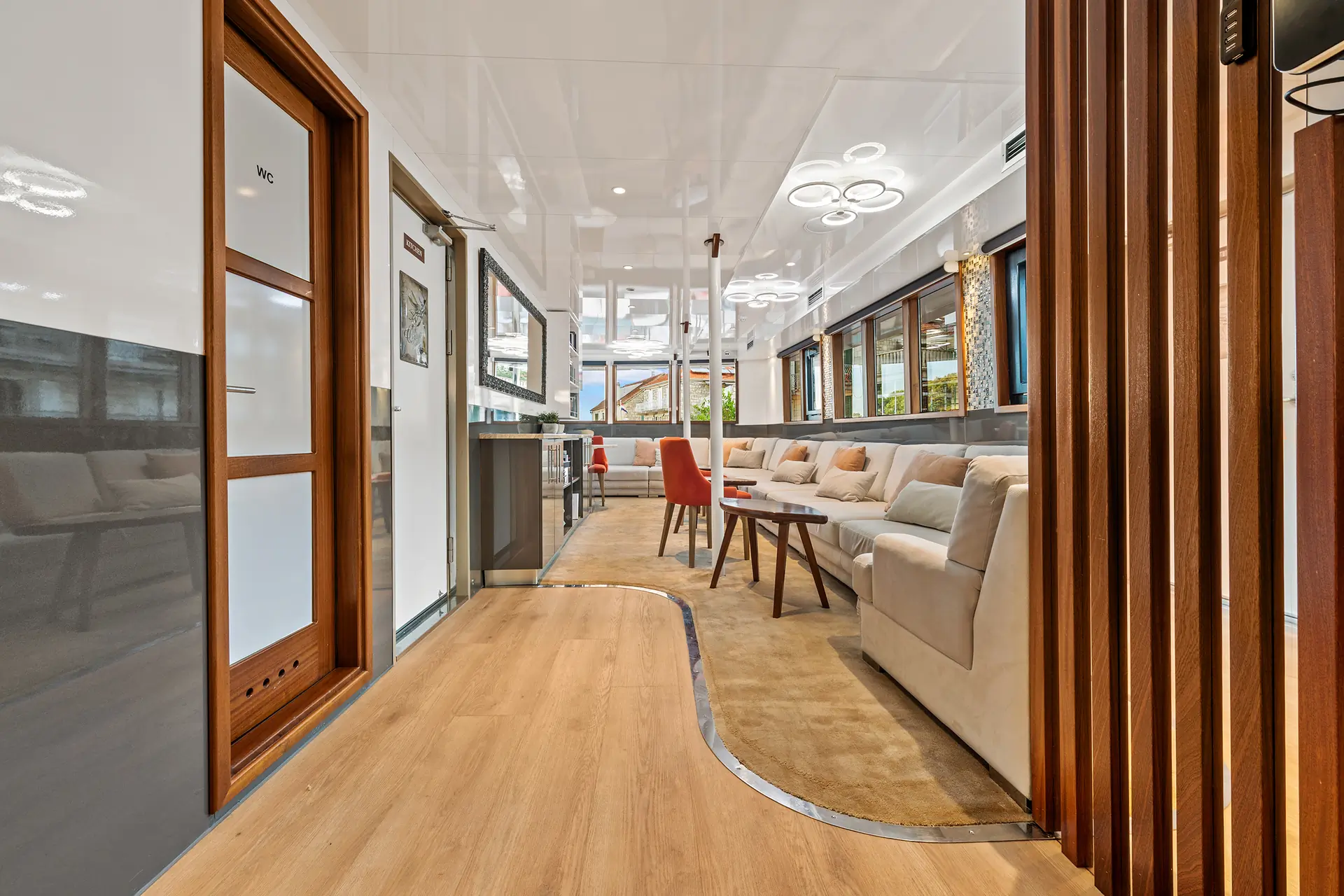 Modern lounge area with wood flooring, beige sofas, round table with chairs, and large windows. Ceiling lights and wood panel accents create a bright, inviting atmosphere.