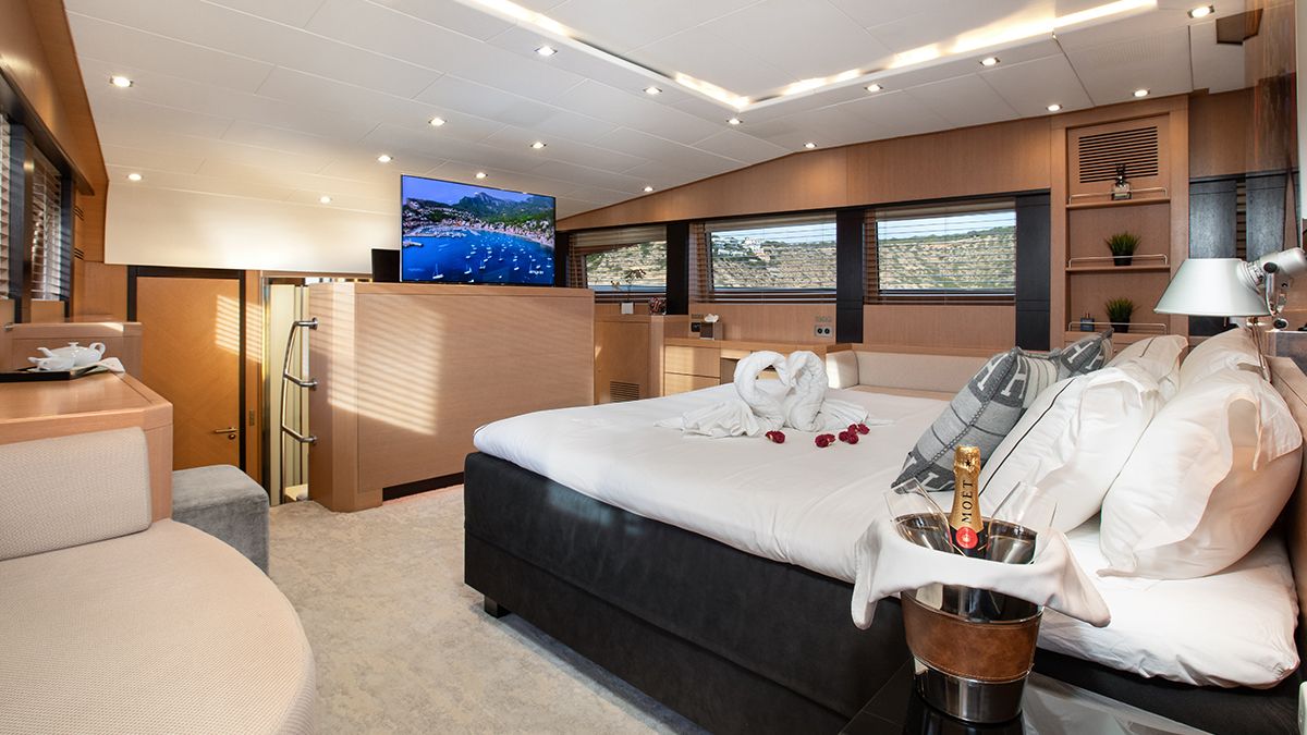 YCM 120 Modern yacht bedroom with a large bed, swan towels and rose petals, TV, windows with ocean view, and champagne on ice beside the bed. Light wood décor and built-in lighting create a luxurious atmosphere.