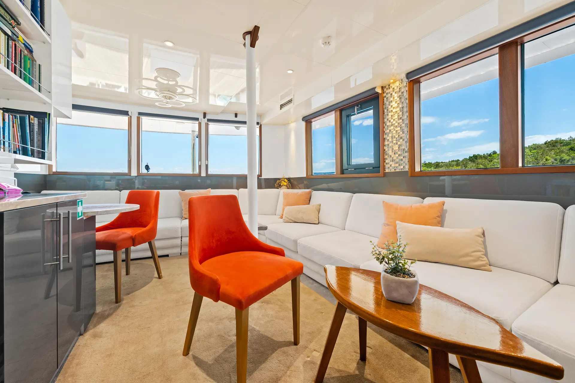 Modern living area with white sofas, orange chairs, a wooden coffee table, bookshelves, and large windows showing a blue sky and greenery outside. A small plant decorates the table.