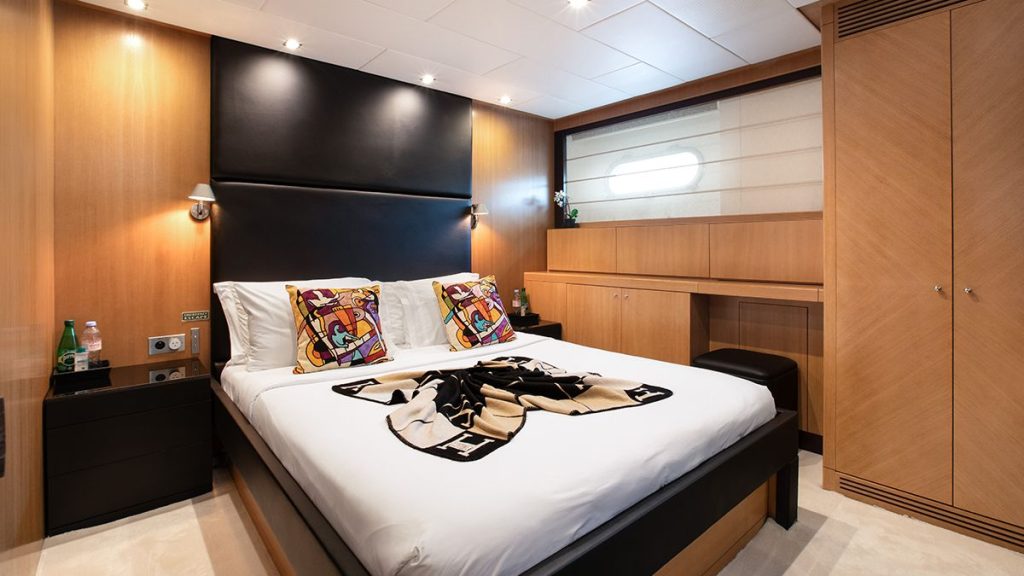 YCM 120 A modern bedroom with a large bed, colorful pillows, a folded blanket, wooden cabinets, a small window, bedside tables with water bottles, wall lights, and neutral décor.