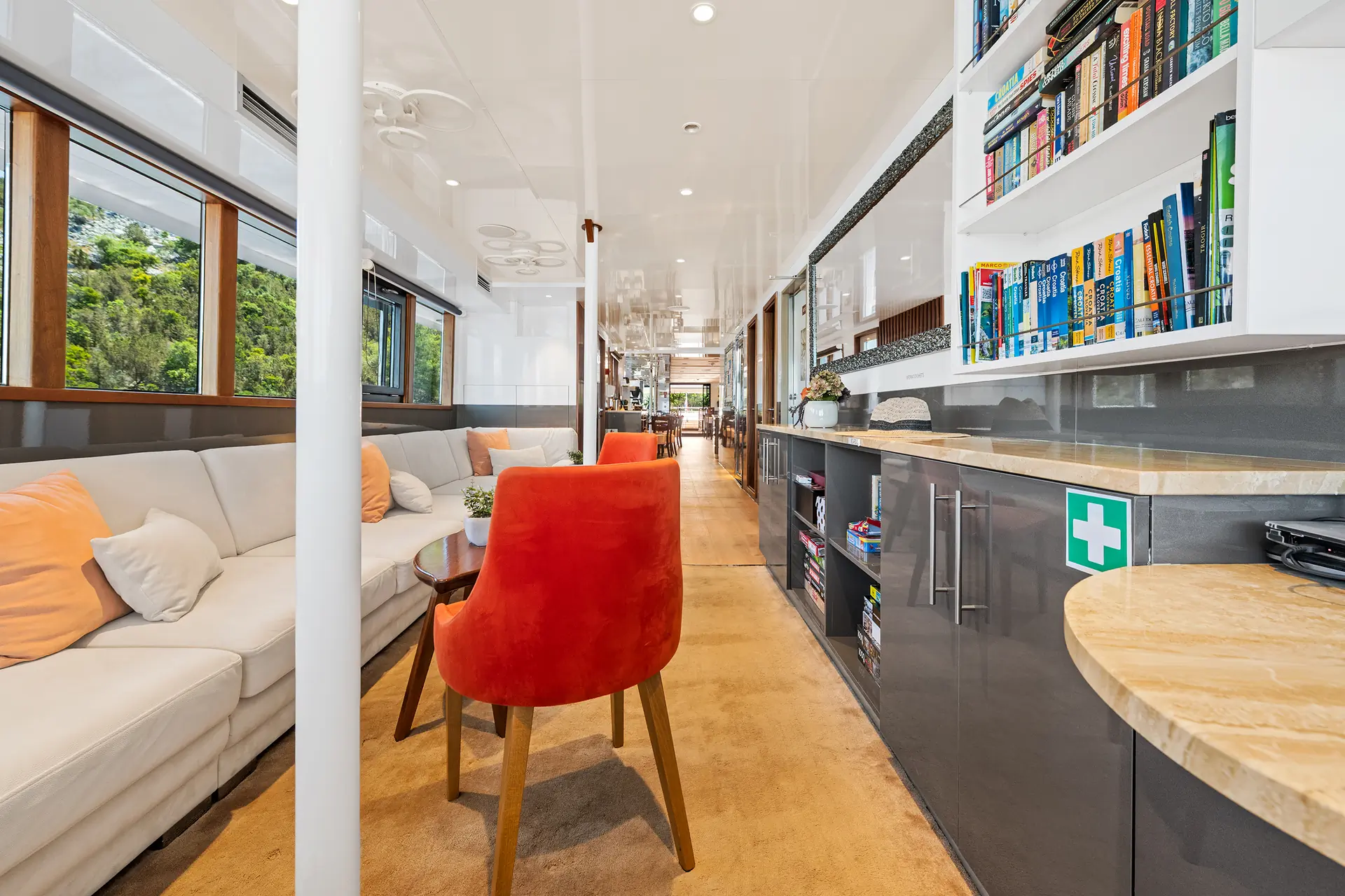 Modern yacht interior with a white sectional sofa, orange chairs, bookshelves, windows with a greenery view, and a first aid kit on the countertop. The space is bright, tidy, and well-lit with ample natural light.