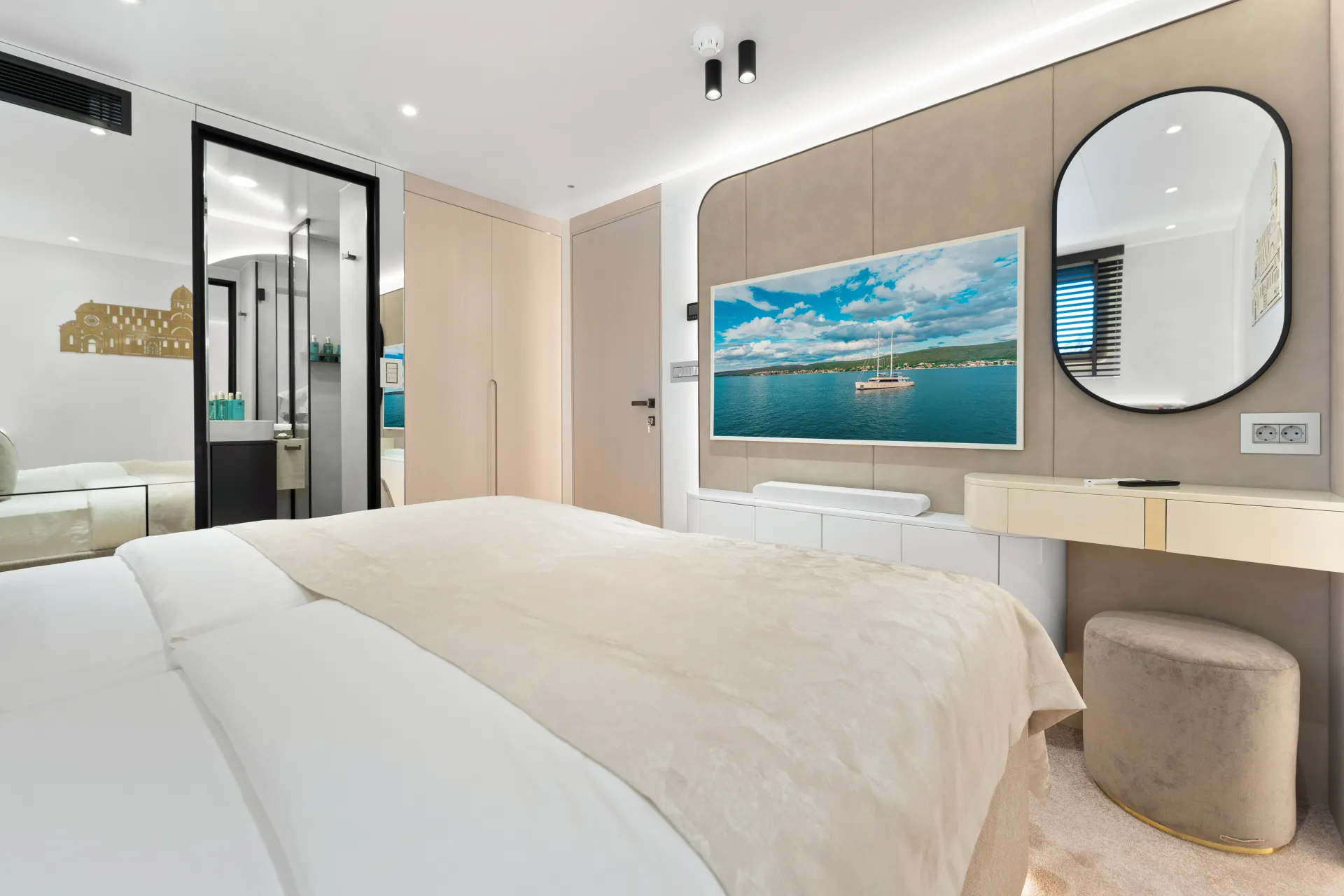 MAXITA Modern bedroom with a large bed, beige bedding, wall-mounted TV displaying a boat on water, round mirror above a vanity table with a cushioned stool, and glass door leading to a bathroom.