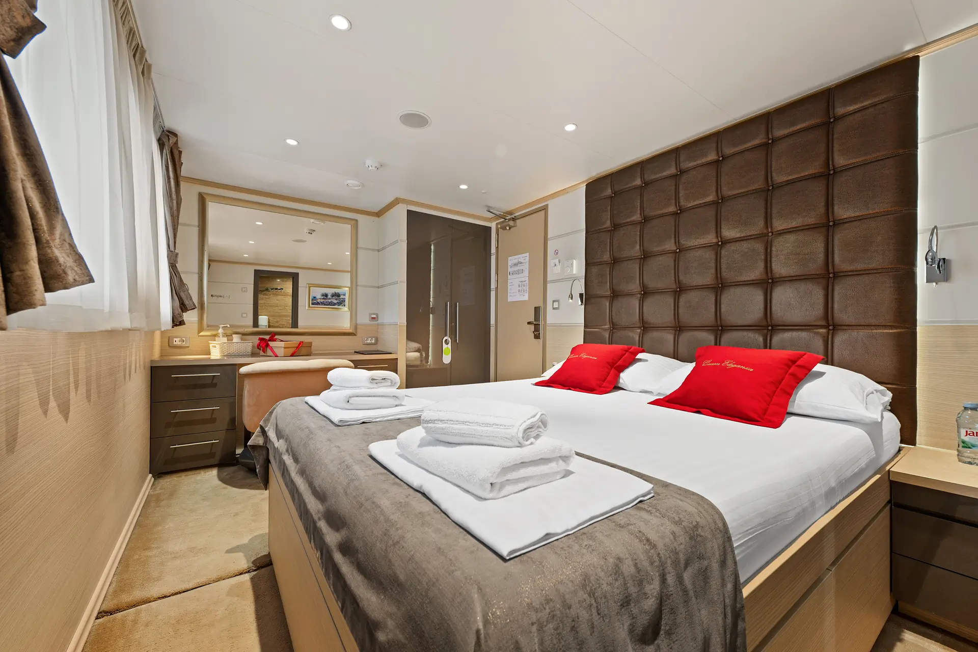 A modern cruise ship cabin with a large double bed, red accent pillows, folded towels, bedside tables, a mirrored vanity, and a window with brown curtains. The room is bright, clean, and neatly arranged.