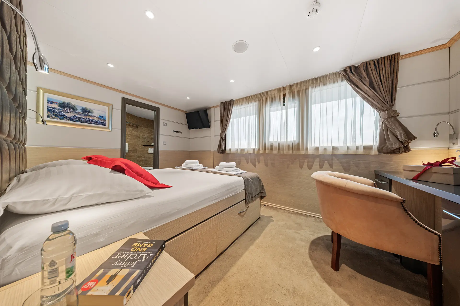 A modern, cozy hotel room with a double bed, desk, chair, TV, large windows with curtains, water bottle, and a book on the table. Towels and red throw pillow are neatly placed on the bed.