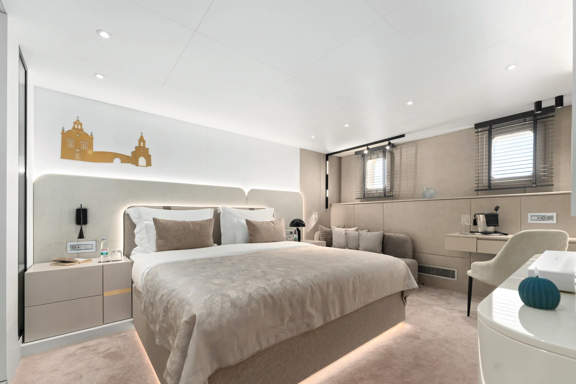 MAXITA Modern hotel room with a large bed, neutral tones, two bedside tables, a sofa by two small windows, and a wall art of a golden skyline. A white desk with a chair and coffee maker is in the corner.