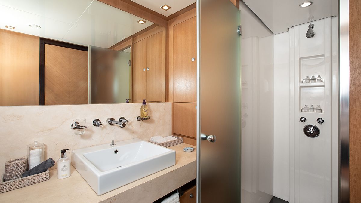 YCM 120 Modern bathroom with a square white sink, wall-mounted faucet, toiletries on the counter, a large mirror, wooden cabinets, and a glass door leading to a shower with shelves and a rainfall showerhead.
