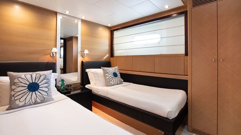 YCM 120 Modern yacht cabin with two single beds, white bedding, and floral pillows. The room features wood-paneled walls, a large mirror, a porthole window, and built-in storage cabinets.