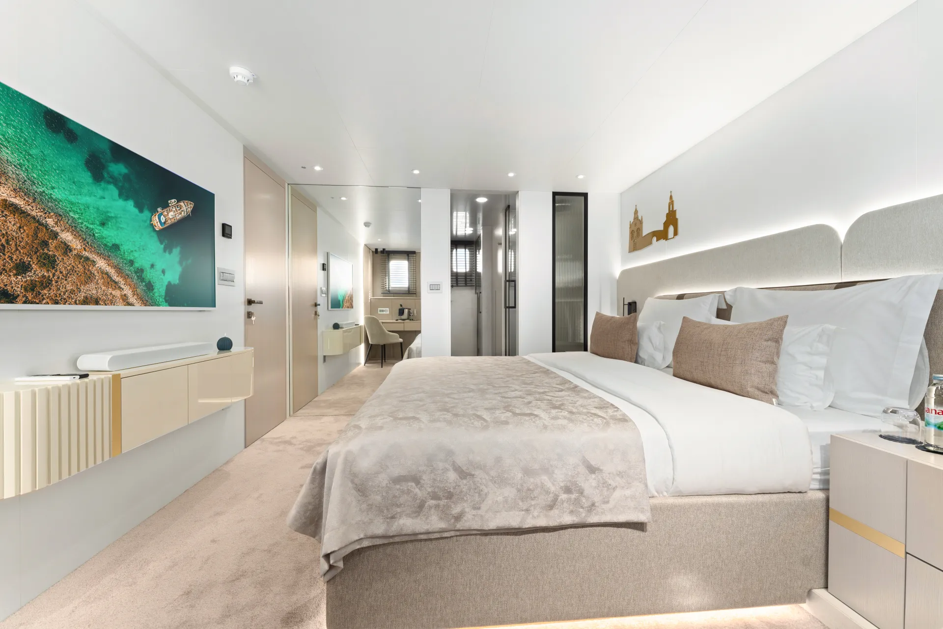 MAXITA Modern hotel room with a large bed, neutral decor, plush pillows, and a beige blanket. A wall-mounted TV displays an aerial view of a boat on water. The room features a mirrored wall, sleek furnishings, and a bright, clean design.