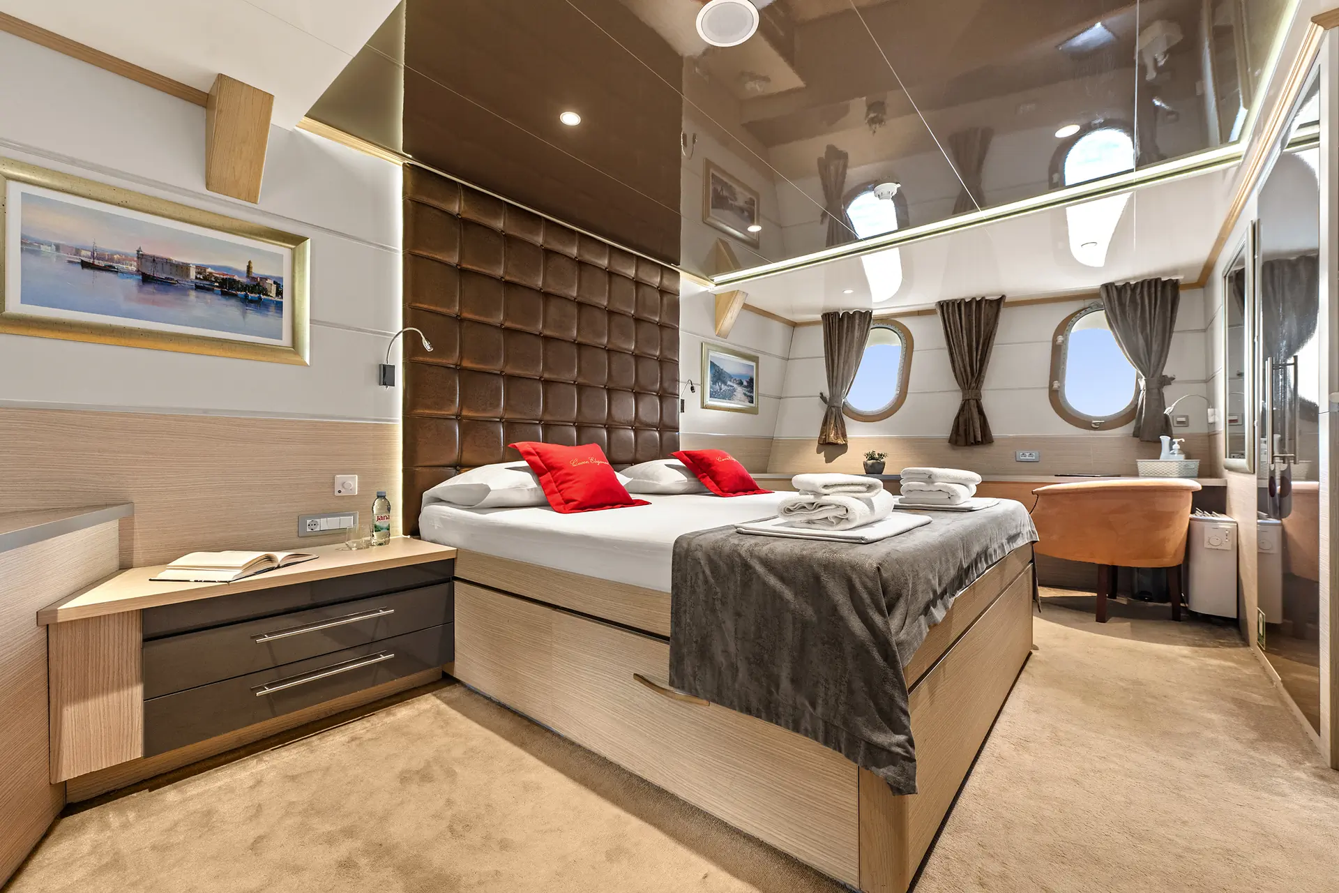 Modern bedroom with a large bed, brown padded headboard, red accent pillows, bedside tables, artwork on the wall, desk by two windows with brown curtains, and folded towels on the bed.